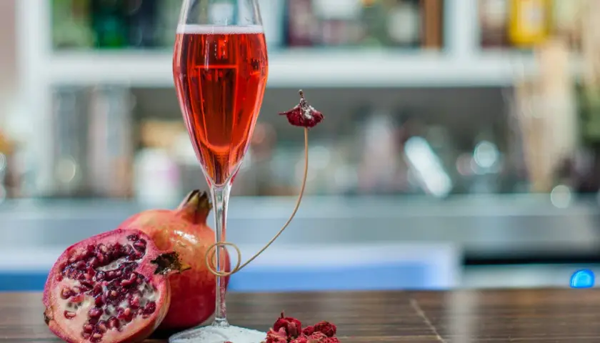 Un bicchiere di tintoretto cocktail rosso rubino, accompagnato da melograni freschi e secchi, su un tavolo di legno scuro.