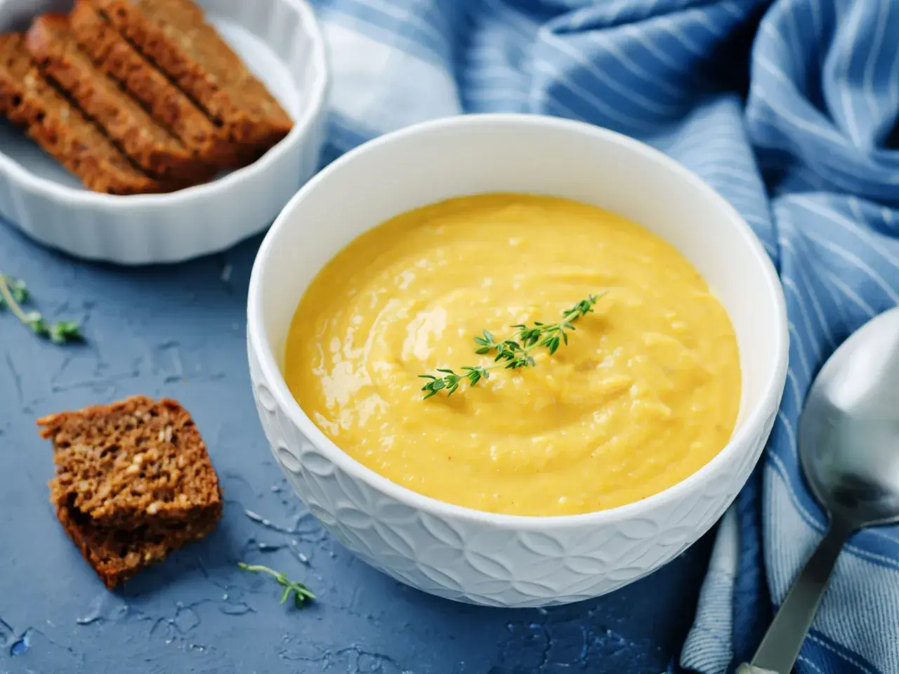 Una ciotola di cremosa salsa di ceci, guarnita con timo, accanto a fette di pane integrale e un tovagliolo blu a righe.