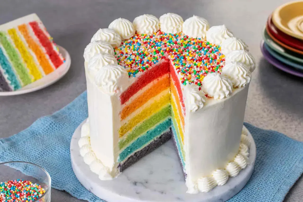 Torta arcobaleno con strati colorati, glassa bianca e zuccherini. Una fetta mostra i colori vivaci.