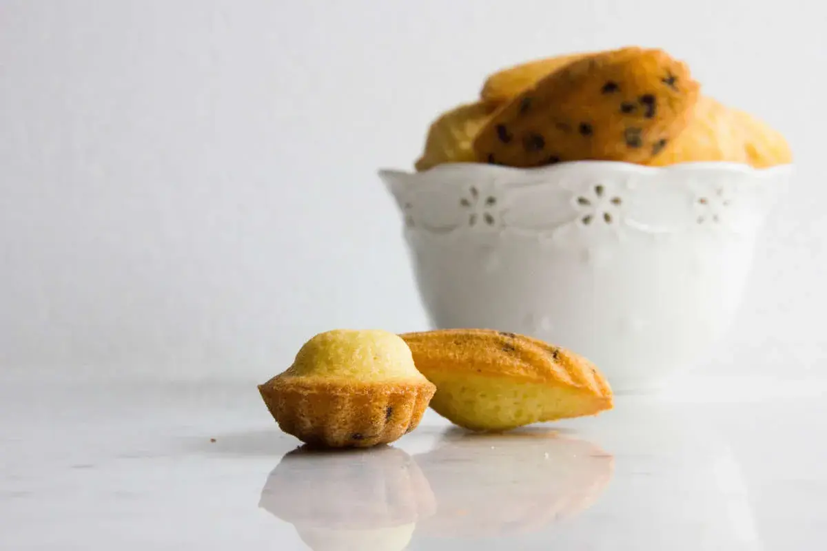 Due madeleine dolci, una a forma di conchiglia e una più tondeggiante, su un piano di marmo. Sullo sfondo, una ciotola bianca piena di altre madeleine.