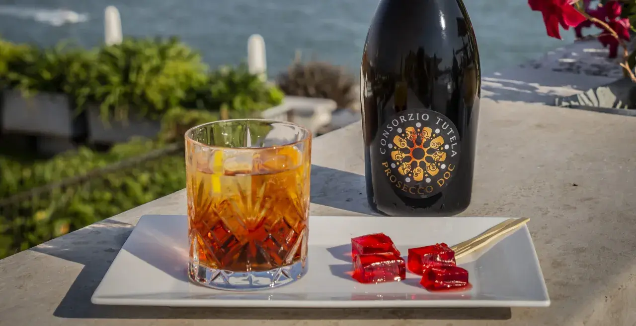 Un bicchiere di spritz con spumante, una bottiglia di Prosecco e cubetti di gelatina rossa su un vassoio bianco, con vista sul mare.