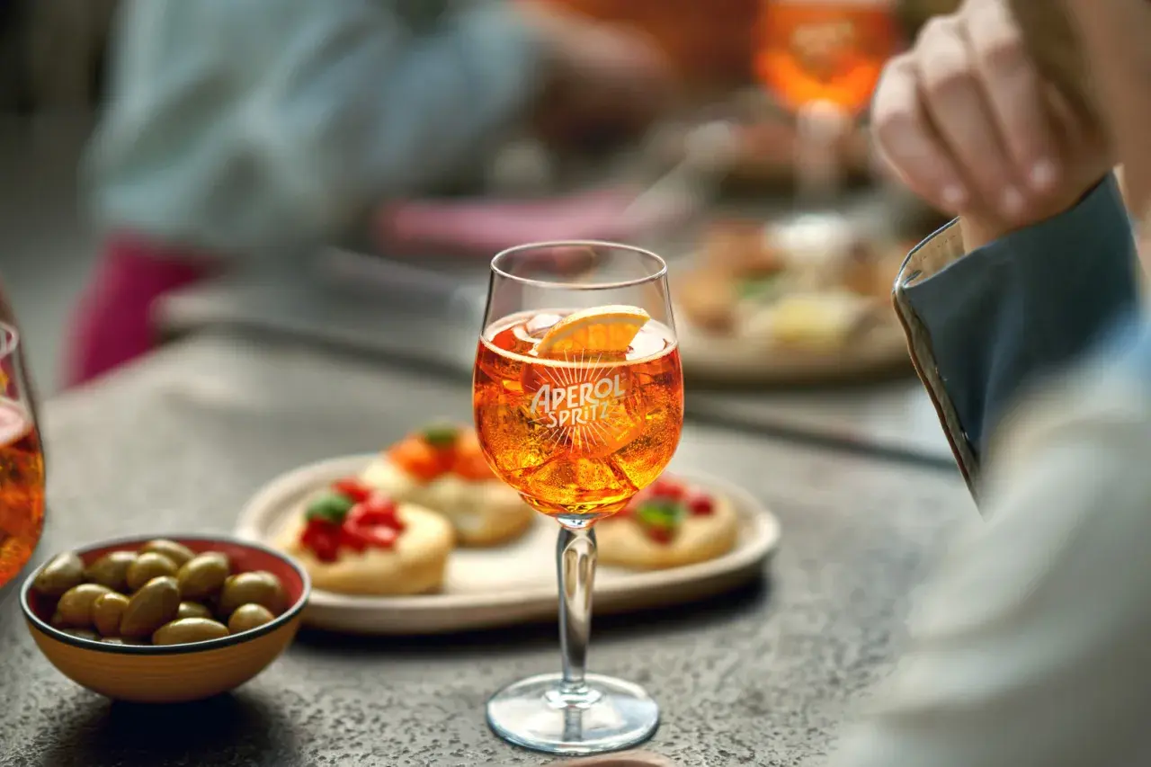 Un bicchiere di Aperol Spritz, ghiaccio e una fetta d'arancia, con olive e stuzzichini sullo sfondo.