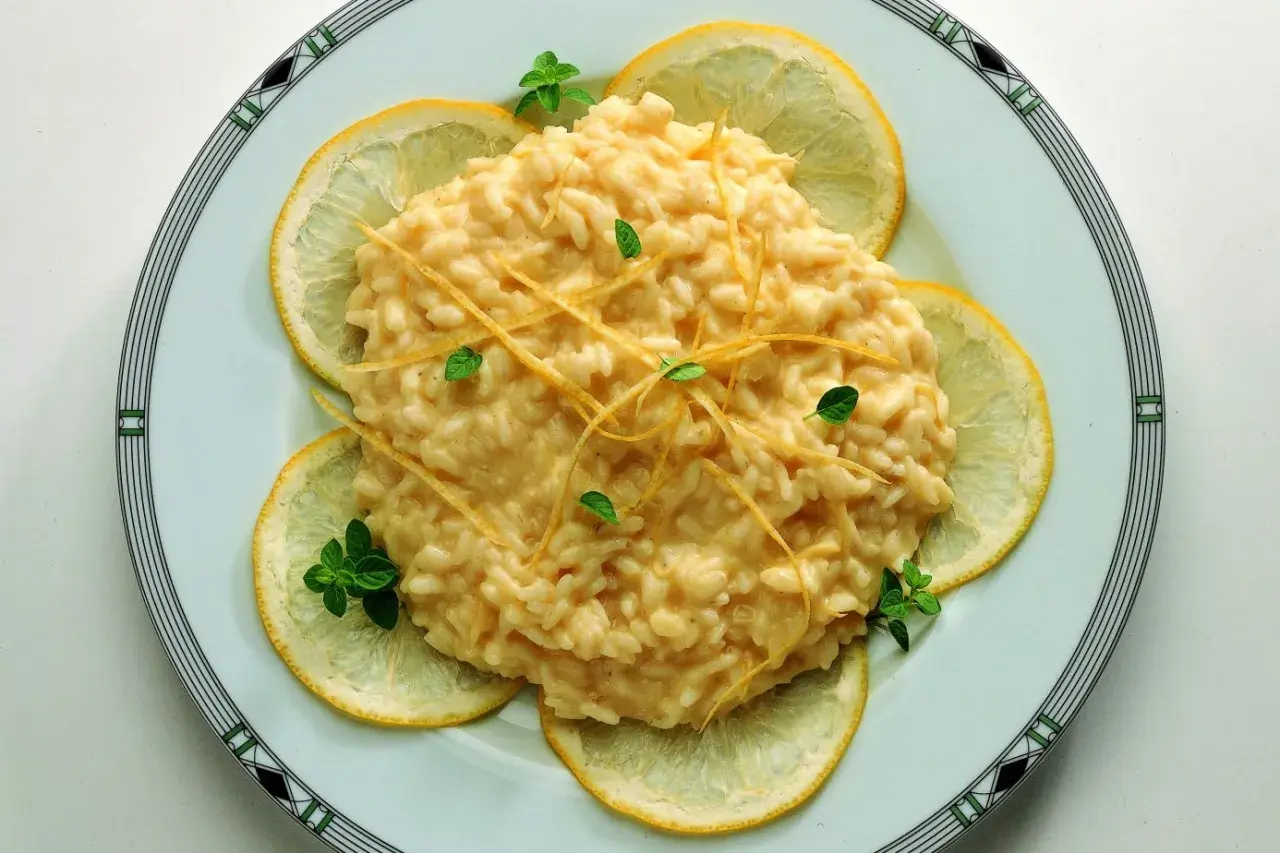 Un cremoso risotto al limone, guarnito con scorzette e foglioline di menta, circondato da fette di limone fresco.