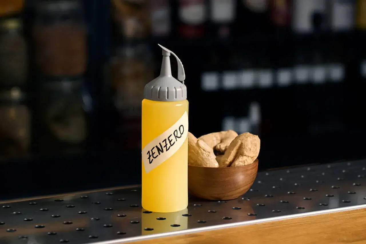 Flacone giallo con etichetta "ZENZERO" e una ciotola di radici di zenzero essiccate.