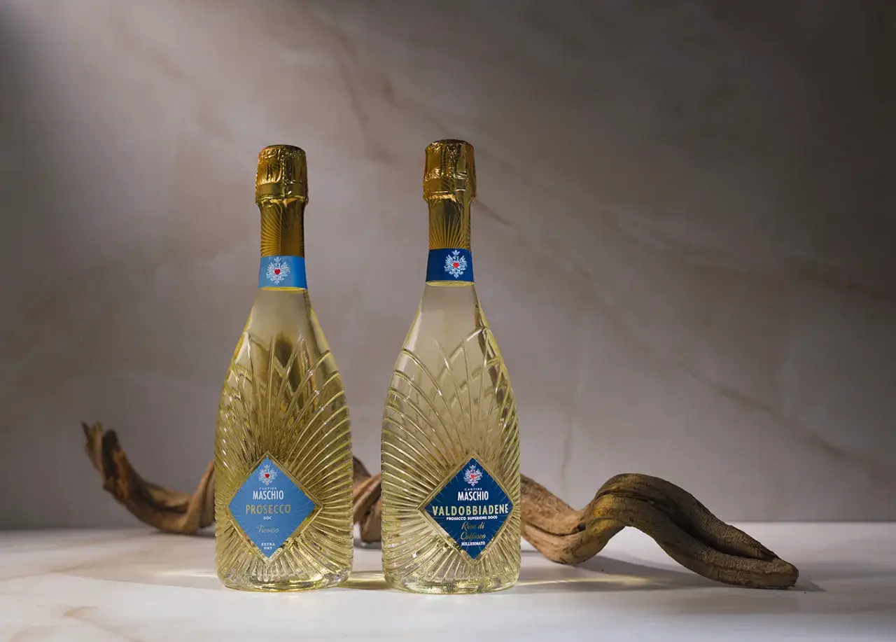 Due bottiglie di Prosecco Maschio, una DOC Treviso e l'altra Valdobbiadene DOCG, con un ramo secco. Scopri come si fa il Prosecco.