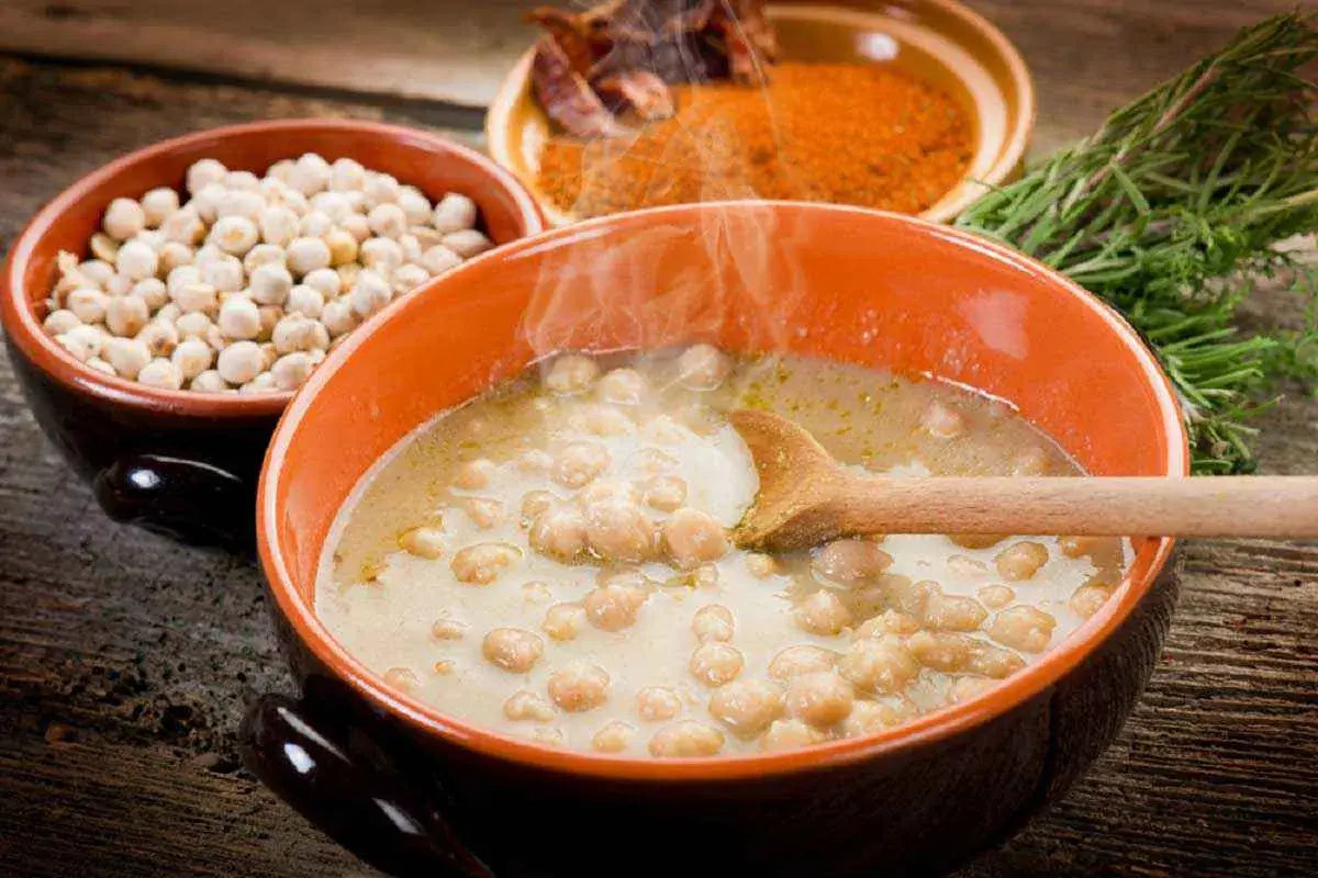 Una fumante zuppa di ceci in una ciotola arancione, con ceci secchi, spezie e rosmarino sullo sfondo.