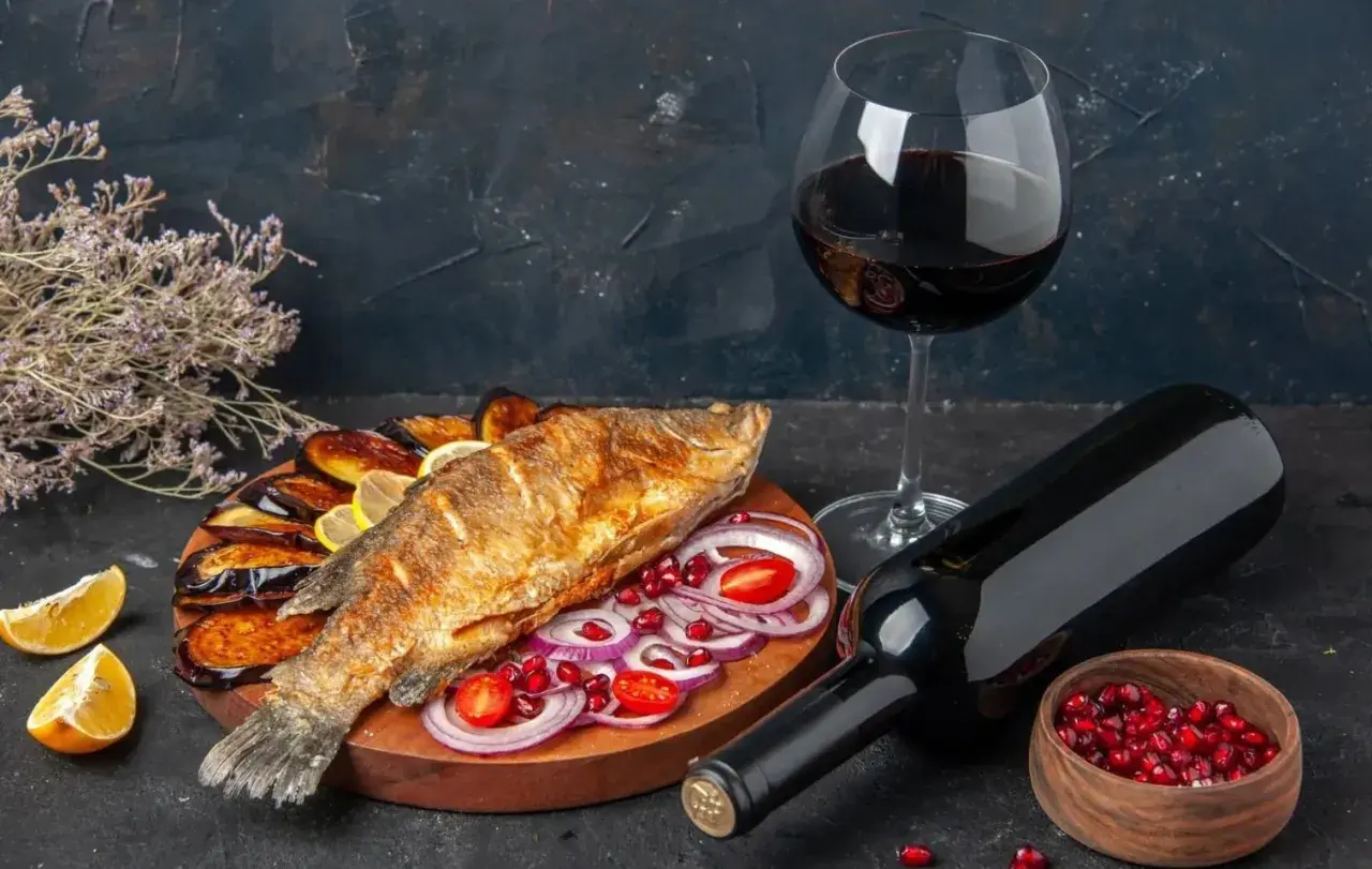 Un delizioso pesce fritto su un piatto con melanzane, cipolle rosse e pomodorini, accompagnato da vino rosso.