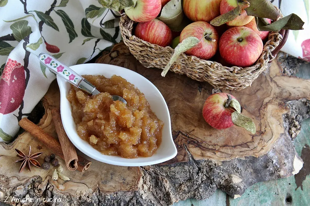 Una ciotola di composta di mele, profumata con cannella e anice stellato, perfetta per la marmellata di natale. Mele rosse in un cesto sullo sfondo.