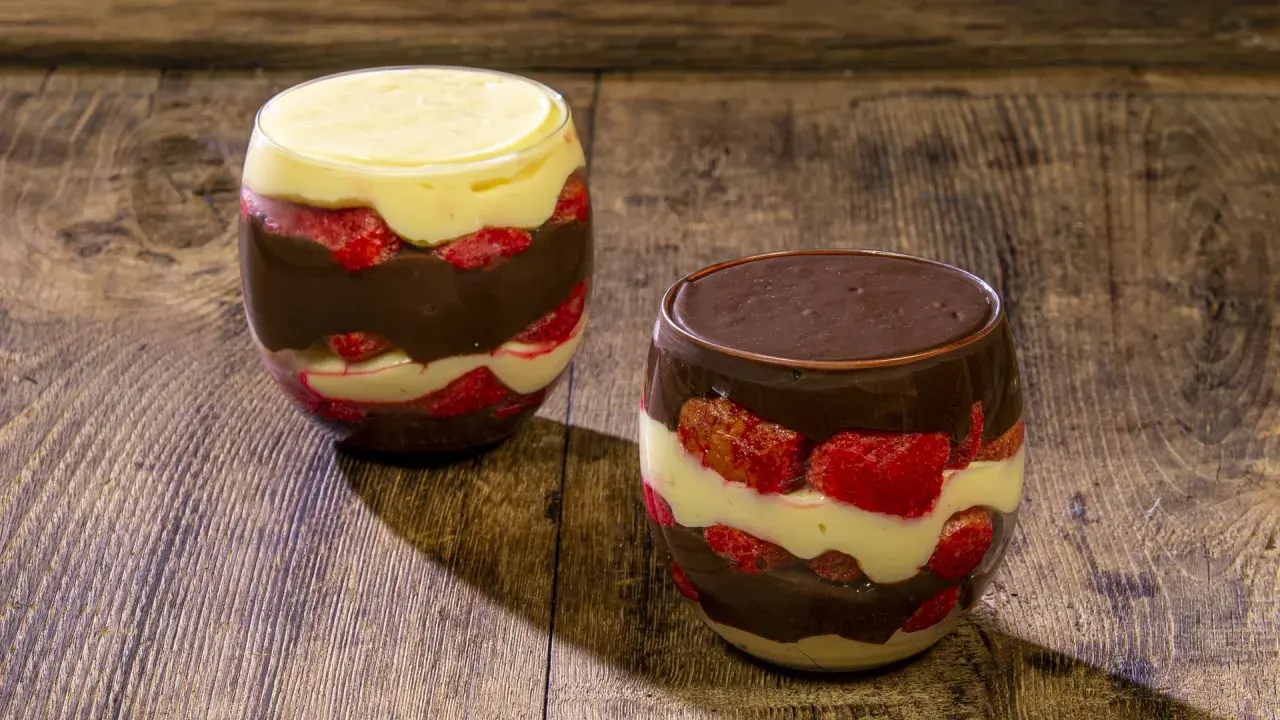 Due bicchieri di zuppa inglese con strati di crema pasticcera, fragole e cioccolato, su un tavolo di legno rustico.