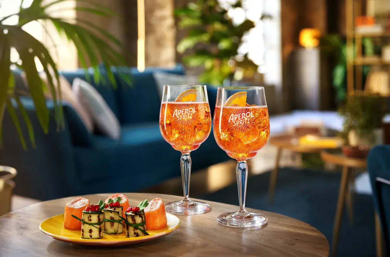 Due bicchieri di cocktail con Aperol Spritz, ghiaccio e fetta d'arancia, accompagnati da stuzzichini su un tavolino in legno.