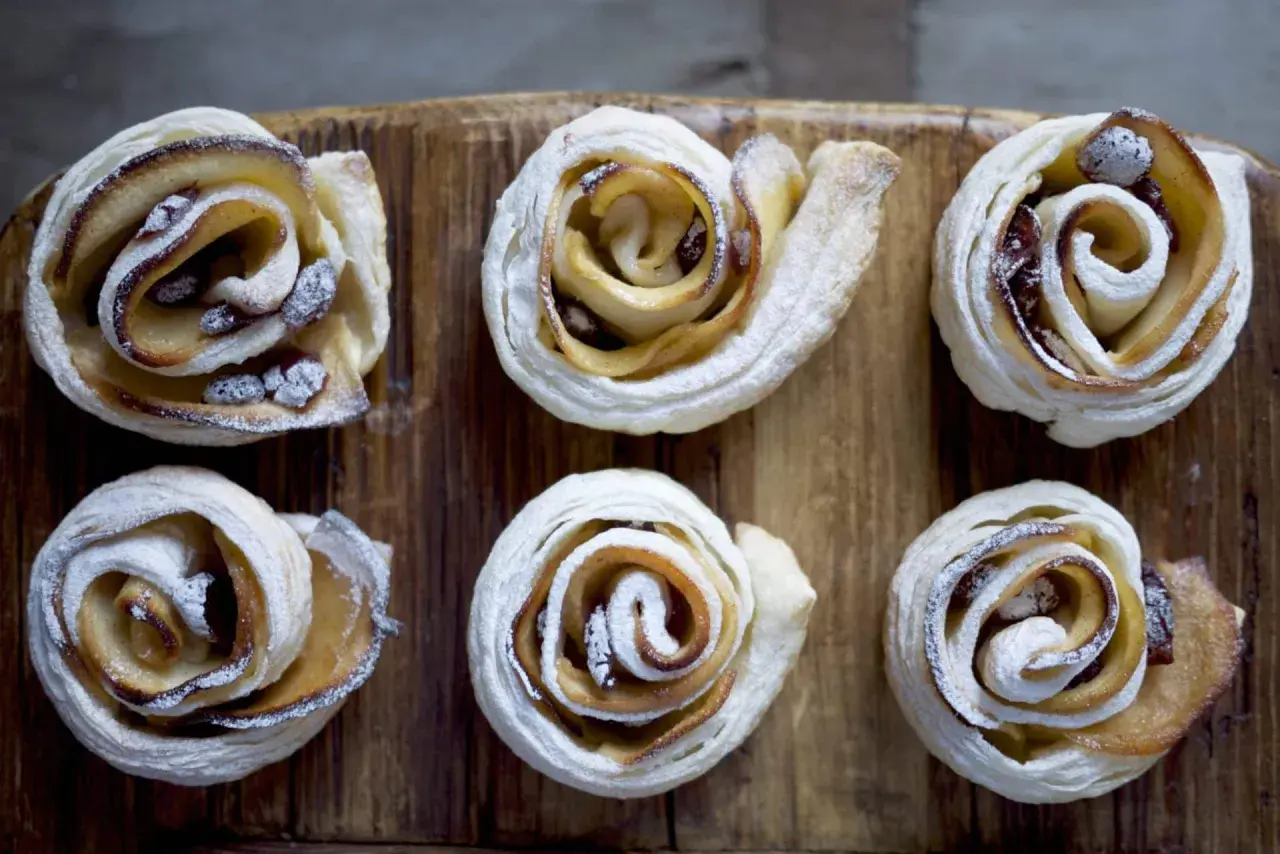 Sei rose di pasta sfoglia con mele e zucchero di canna, cosparse di zucchero a velo, su un tagliere di legno.