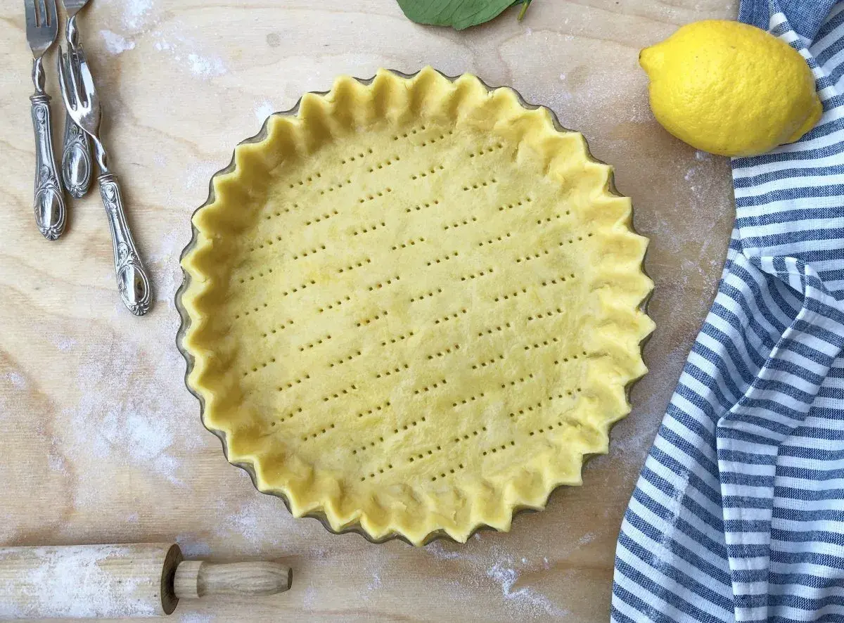 Base di pasta frolla pronta in teglia, forata e con bordi ondulati, accanto a un limone e un canovaccio a righe.