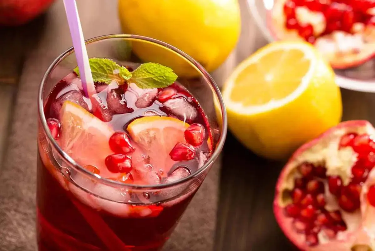 Un rinfrescante spritz al melograno con fette di limone, chicchi rossi e foglie di menta, accompagnato da limoni e melograni freschi.