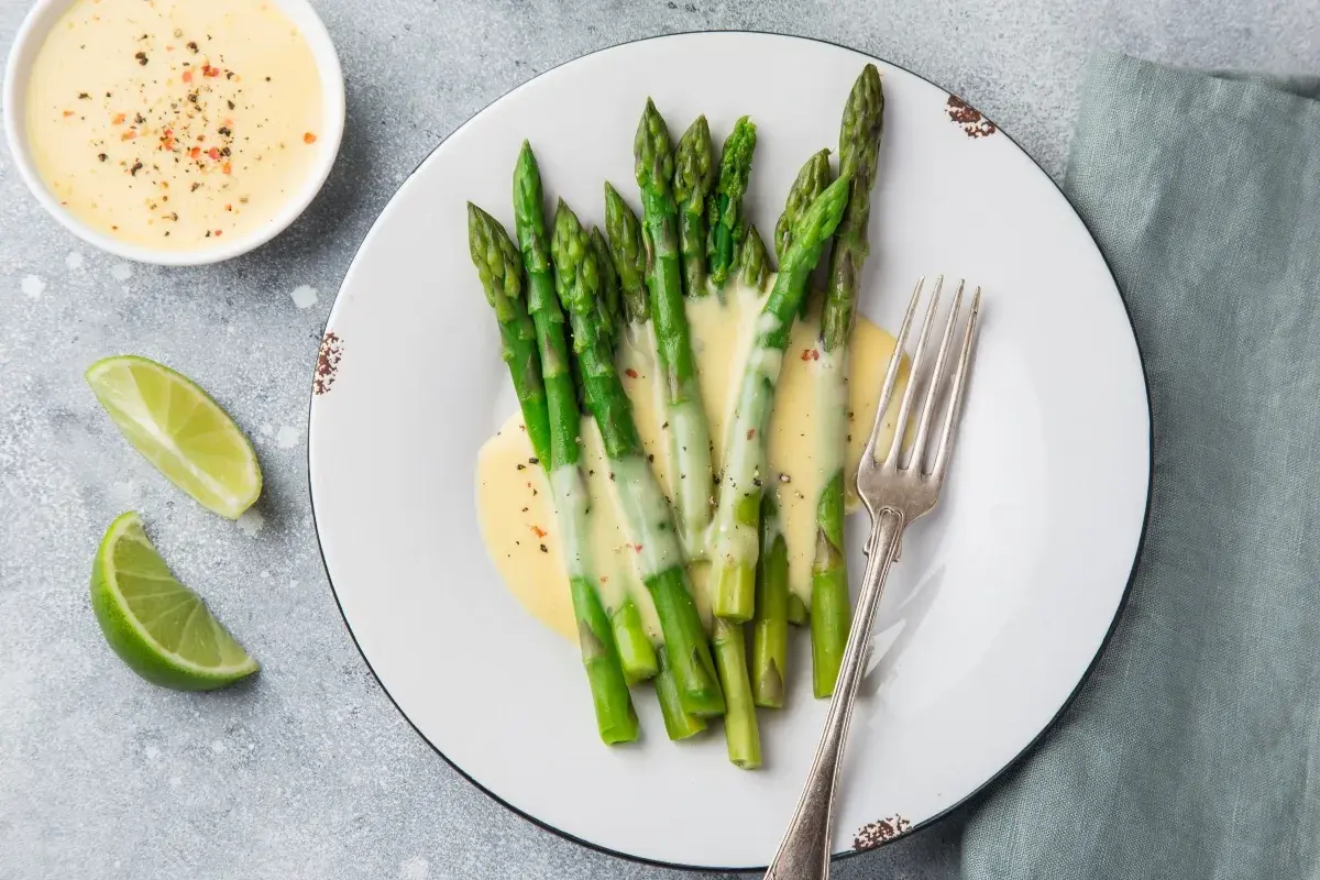 Asparagi freschi conditi con una cremosa salsa olandese, accompagnati da spicchi di lime e una forchetta d'argento.