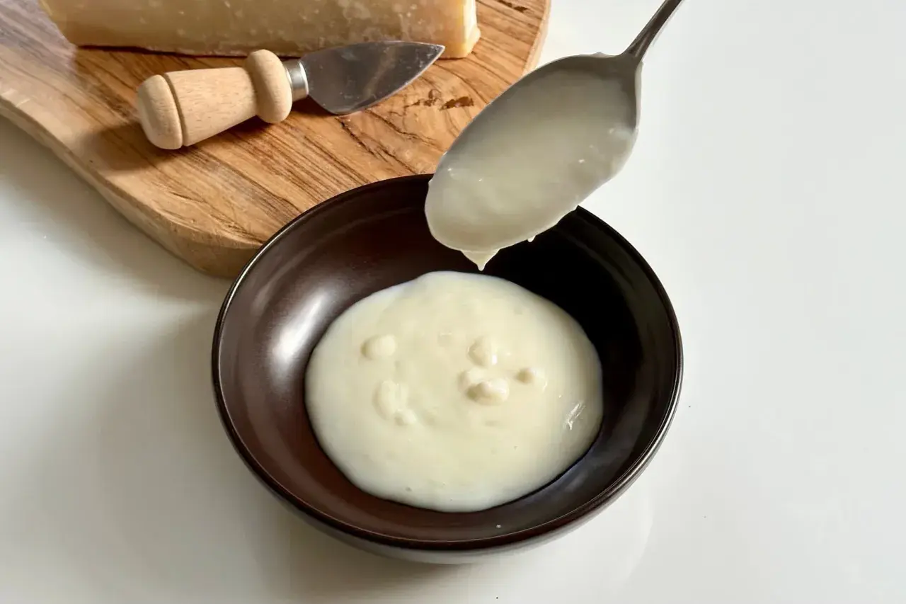 Un cucchiaio versa una densa crema di parmigiano in una ciotola. Sul tagliere, un pezzo di formaggio e un coltellino.