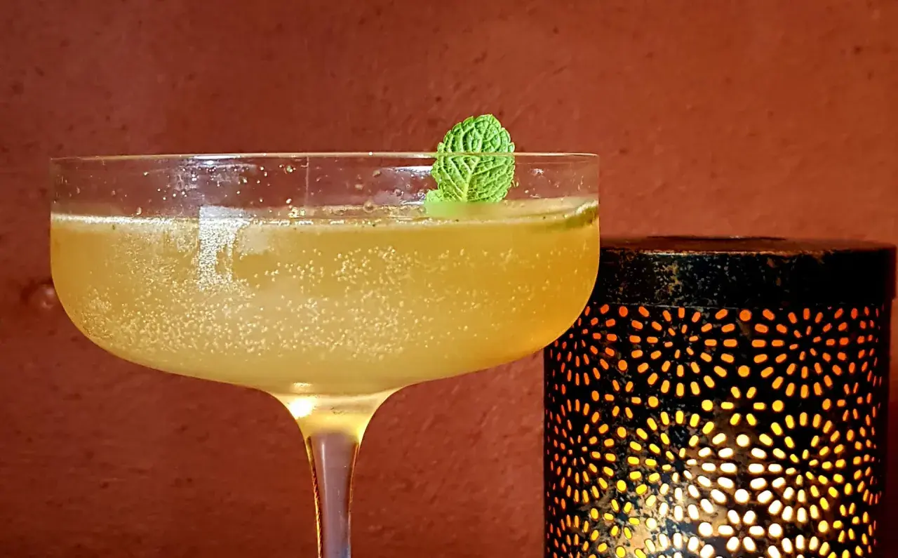Rinfrescante cocktail con rum e menta, guarnito con una fogliolina verde, accanto a una lanterna decorativa che emana luce calda.