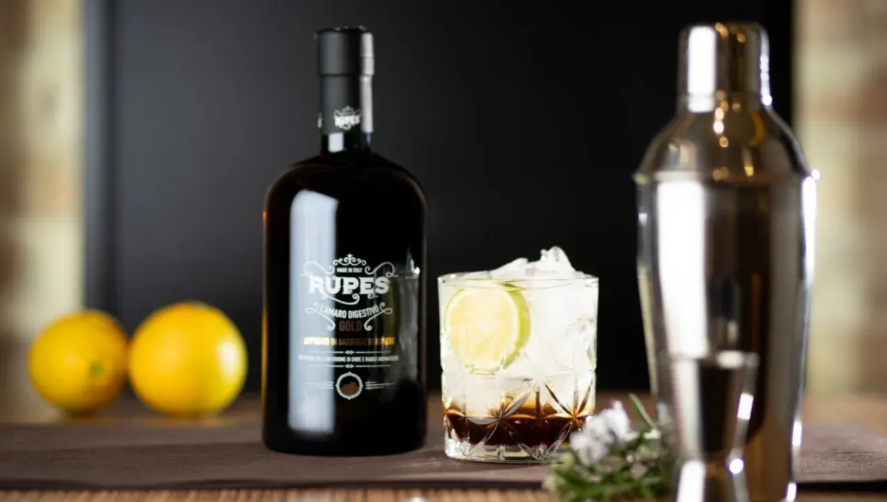 Bottiglia di amaro Rupes, un classico tra gli amari calabresi, accanto a un bicchiere con ghiaccio, lime e un cocktail.