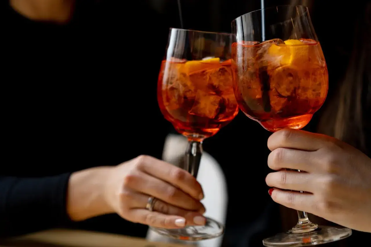 Due mani brindano con due spritz, un superalcolico rinfrescante con ghiaccio e arancia, su sfondo scuro.