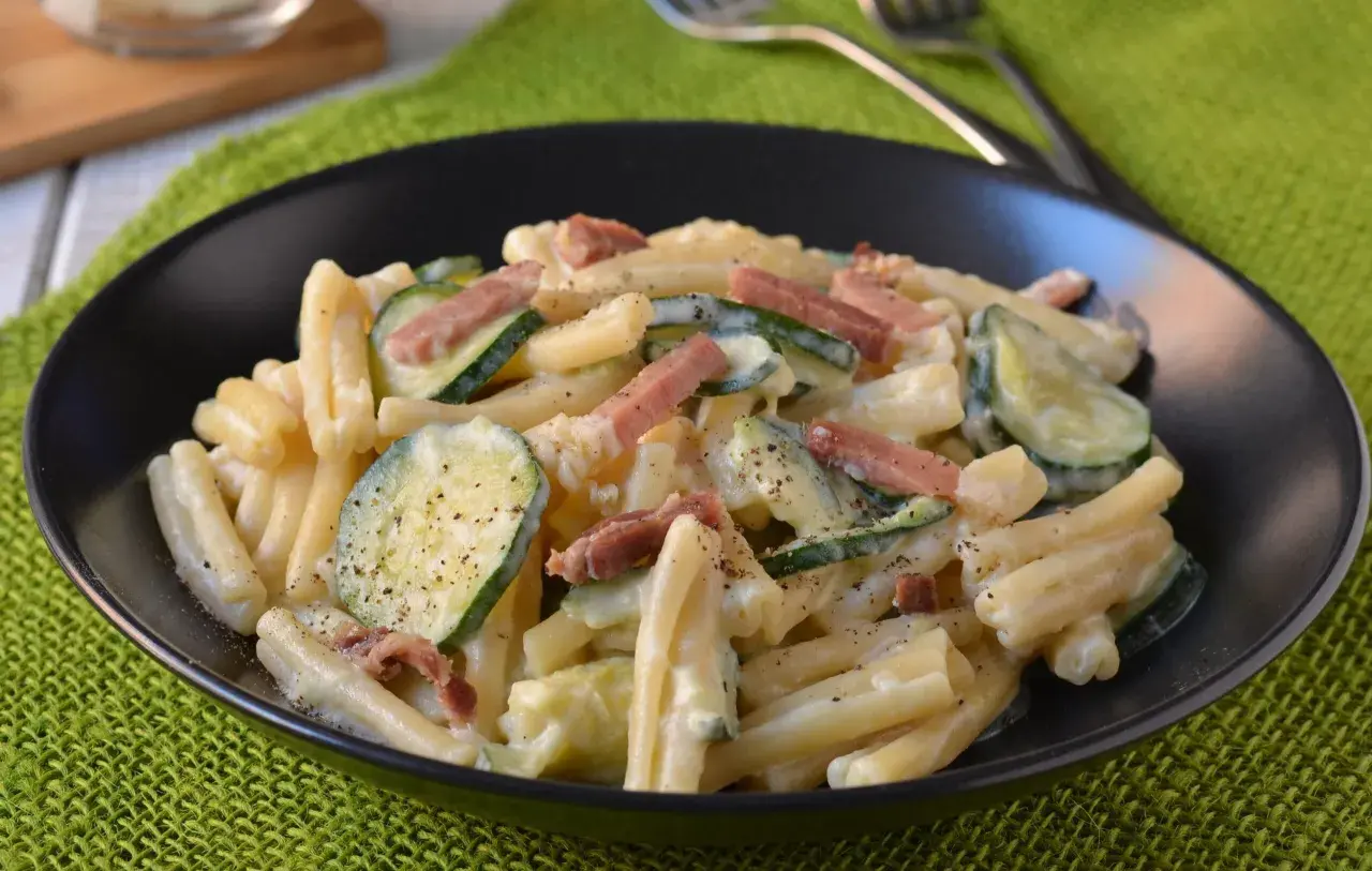 Piatto di pasta cremosa con zucchine a rondelle e cubetti di speck croccante, guarnito con pepe nero.