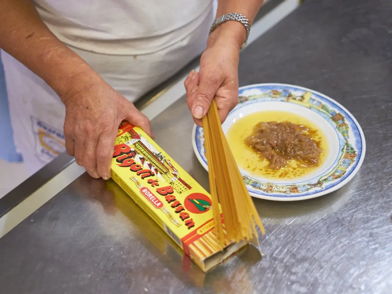 Mani che preparano i bigoli in salsa, con la scatola di pasta e un piatto di salsa di acciughe e cipolle.