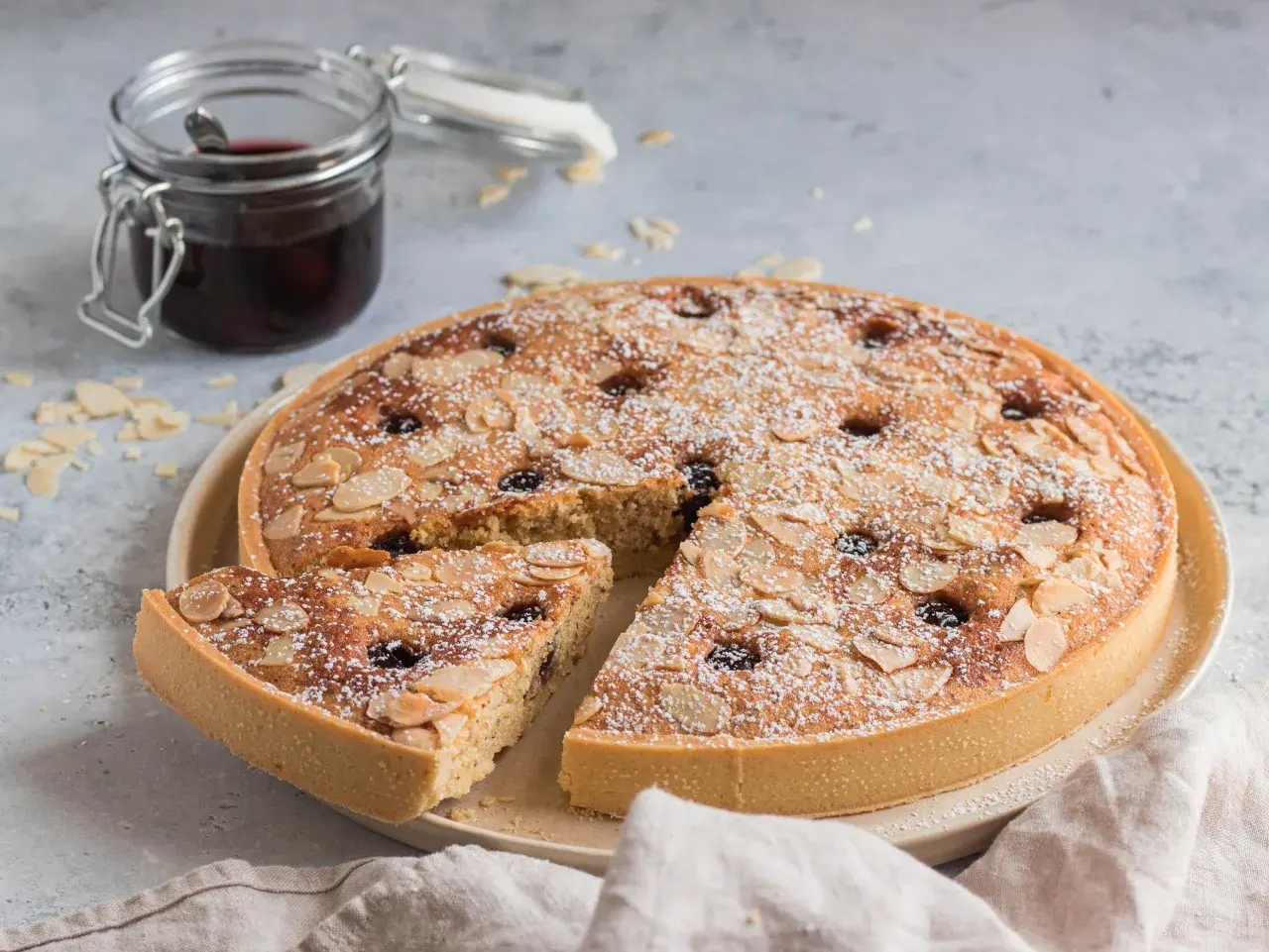 Torta con crema frangipane, mandorle a lamelle e ciliegie, spolverata di zucchero a velo. Accanto, un vasetto di confettura.