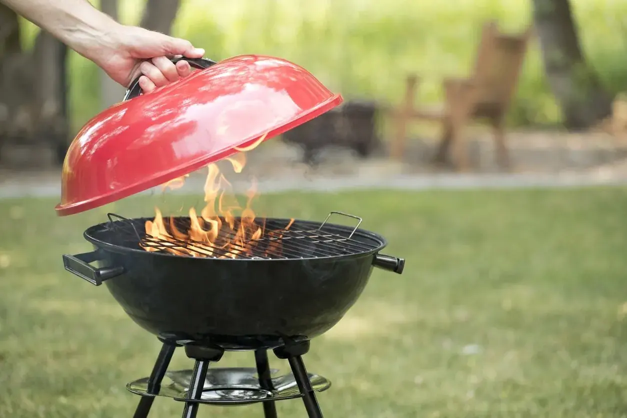 Mano solleva il coperchio rosso di un barbecue a carbonella con fiamme vive. Perfetto per chi vuole sapere se posso usare legna nel barbecue a carbonella.