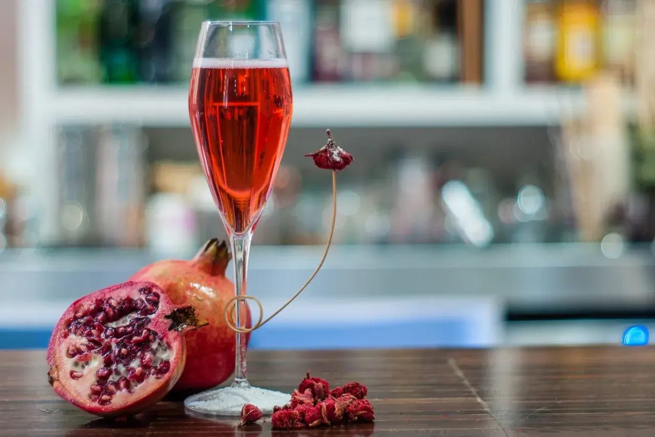 Un bicchiere di tintoretto cocktail rosso rubino, accompagnato da melograni freschi e secchi, su un tavolo di legno scuro.