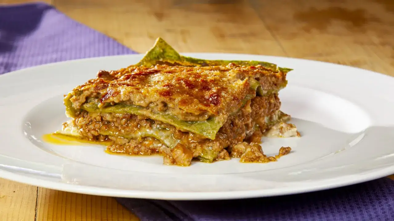 Una porzione di lasagne alla bolognese, con strati di pasta verde, ragù e formaggio fuso, servita su un piatto bianco.
