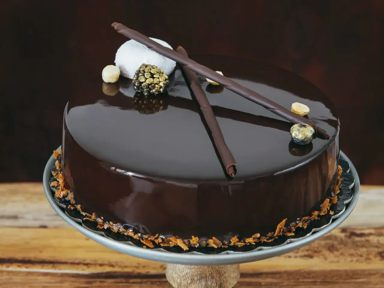 Torta al cioccolato con glassa a specchio facile, decorata con riccioli di cioccolato, nocciole e una sfera dorata.