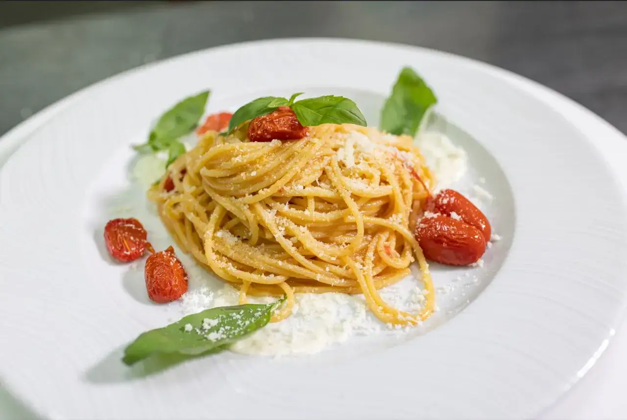 Un piatto di pasta allo scarpariello, spaghetti conditi con pomodorini confit, basilico fresco e abbondante parmigiano grattugiato su un elegante piatto bianco.