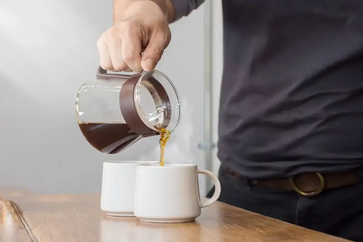 Un uomo versa caffè da una caraffa in due tazze bianche. La ricetta per un caffè americano perfetto inizia così.