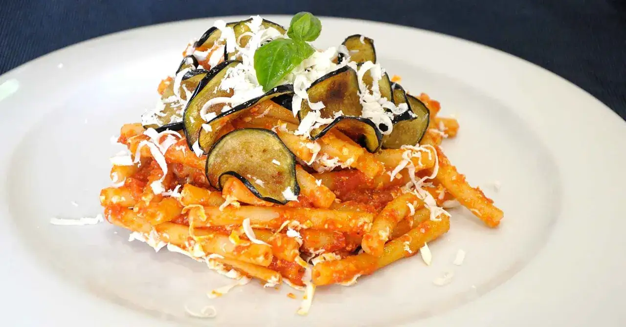 Un piatto di pasta alla norma con melanzane fritte, pomodoro e ricotta salata grattugiata, guarnito con basilico fresco.