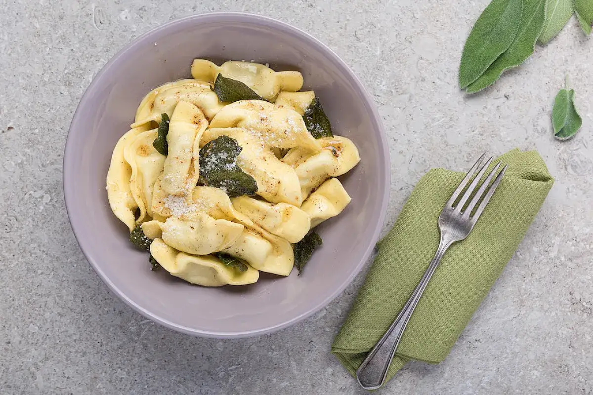 Casoncelli alla bergamasca fumanti in una ciotola, conditi con salvia e parmigiano. Accanto, un tovagliolo verde e una forchetta.