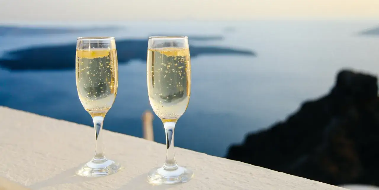 Due calici di spumante, forse Prosecco, con bollicine e una fetta di limone, su uno sfondo marino al tramonto.