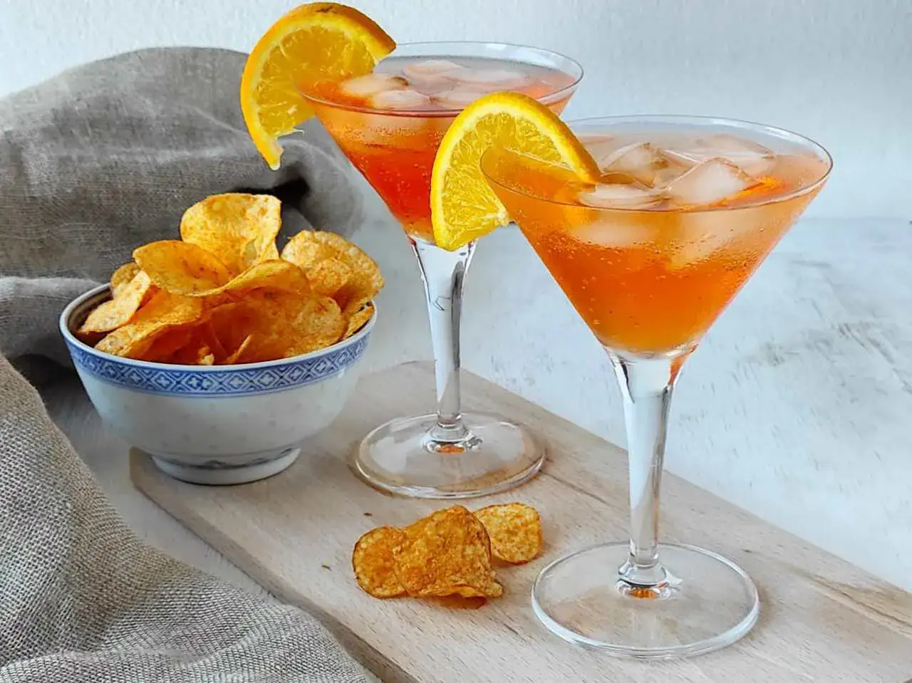 Due spritz con spumante, ghiaccio e arancia, accompagnati da una ciotola di patatine croccanti.