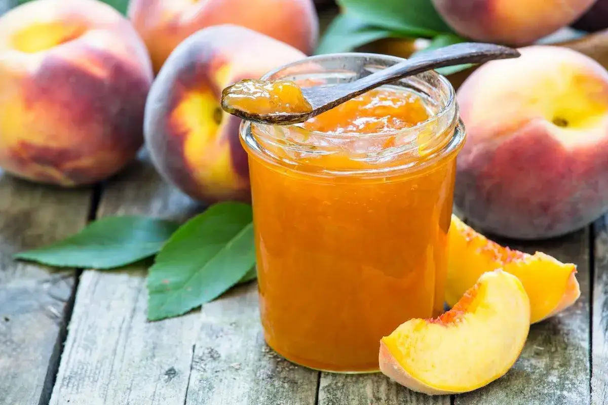 Barattolo di marmellata di pesche su un tavolo di legno, con pesche fresche e foglie verdi intorno. Un cucchiaino raccoglie un po' di marmellata.