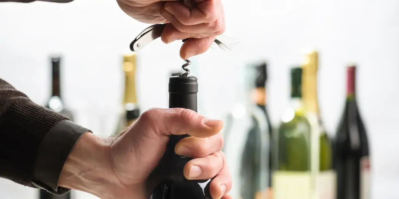 Mani che usano un cavatappi per aprire una bottiglia di vino, con altre bottiglie sullo sfondo.