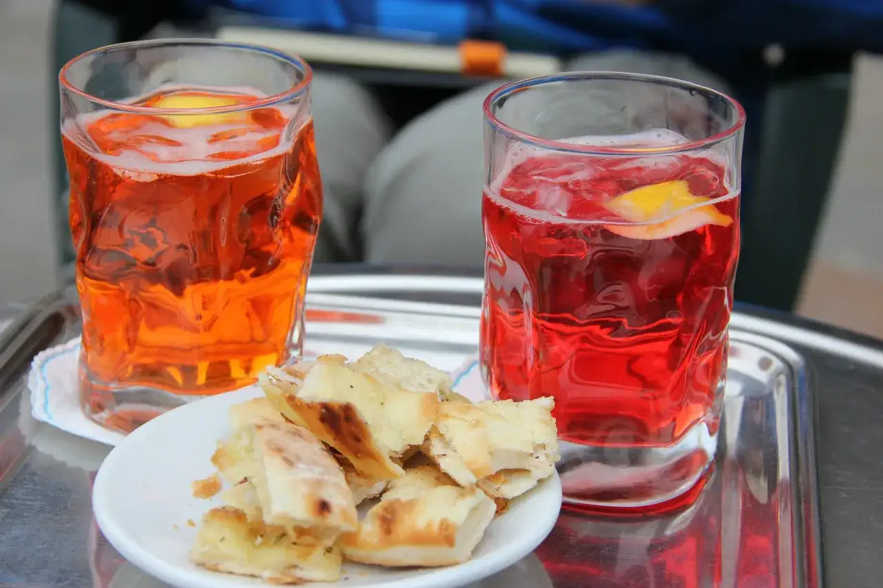 Due bicchieri di spritz aperitivo con amici, uno arancione e uno rosso, accompagnati da focaccia su un tavolino.