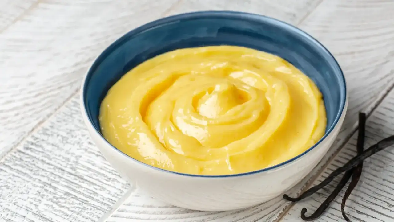 Crema pasticcera liscia e vellutata, preparata con amido di mais, servita in una ciotola blu. Accanto, baccelli di vaniglia.