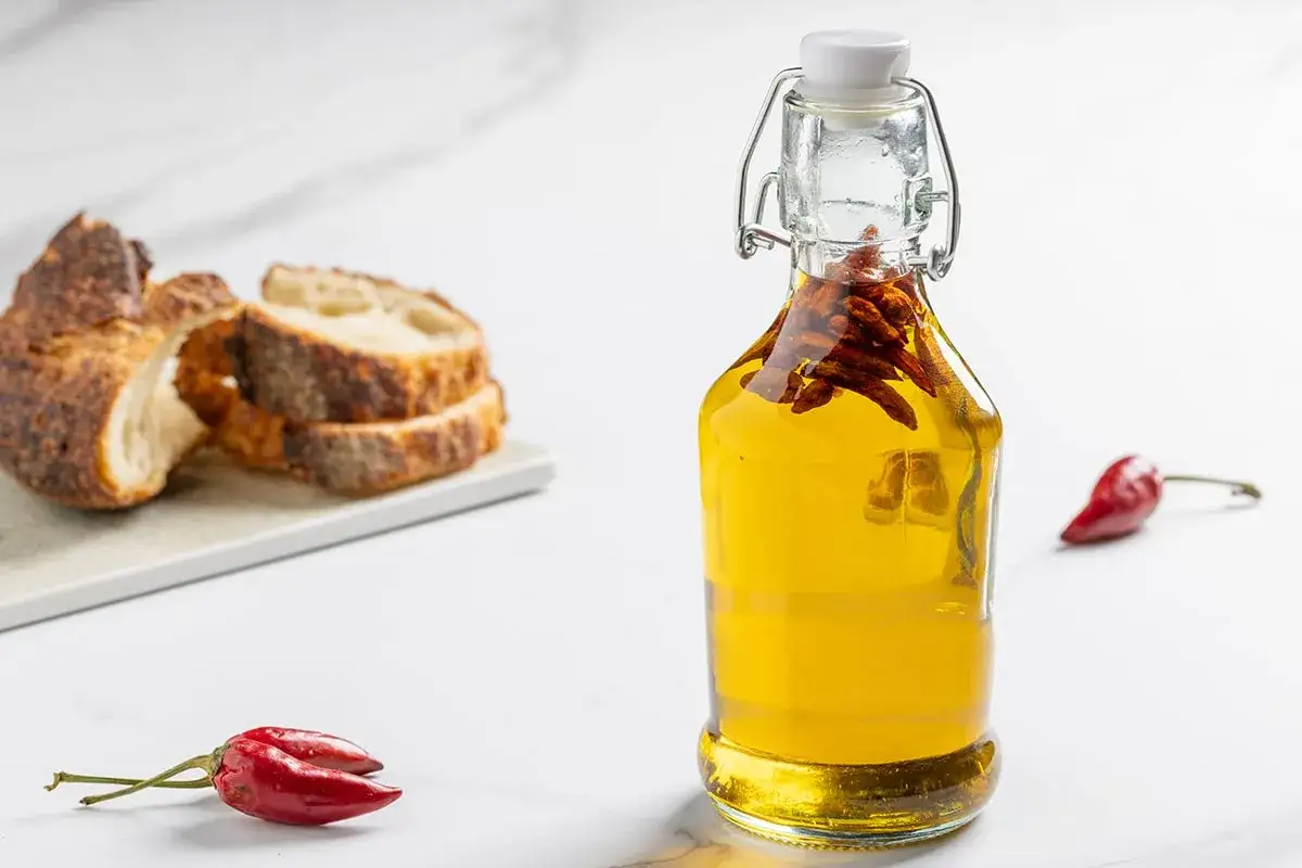 Olio al peperoncino in bottiglia, con peperoncini secchi e pane rustico affettato sullo sfondo.