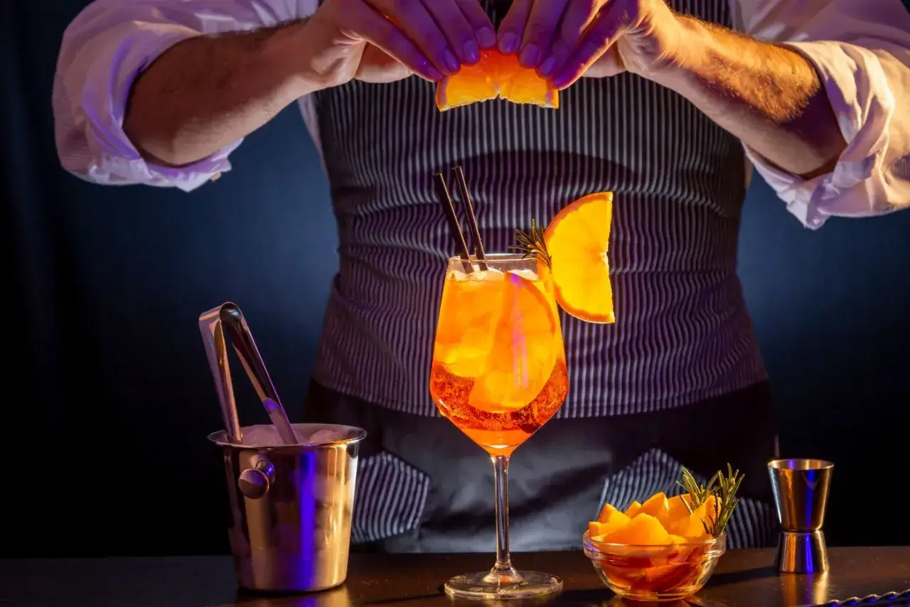 Un barista prepara un Aperol Spritz, il drink simbolo di spritz Italia, guarnendolo con fette d'arancia e rosmarino.