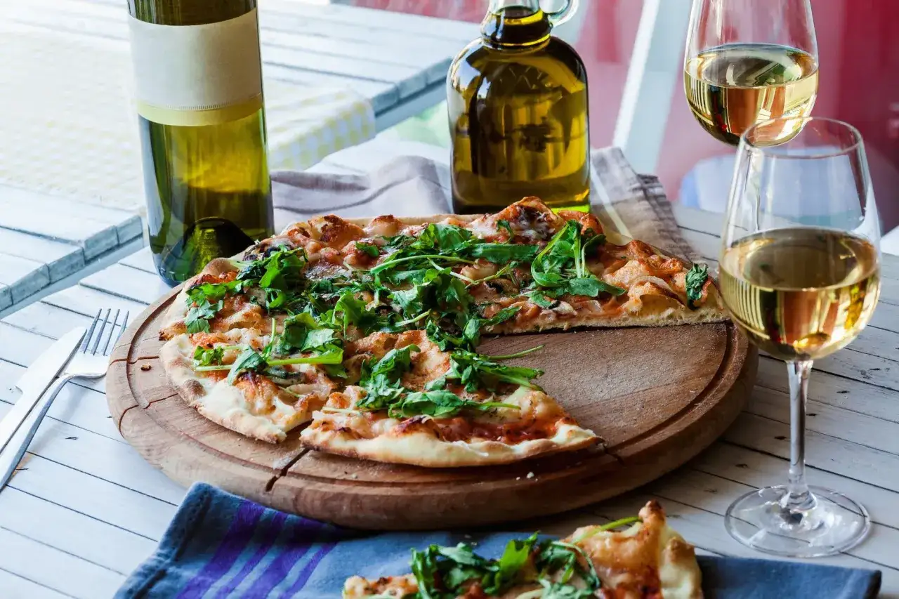 Pizza con cipolla e rucola su tagliere, accompagnata da vino bianco e olio d'oliva.
