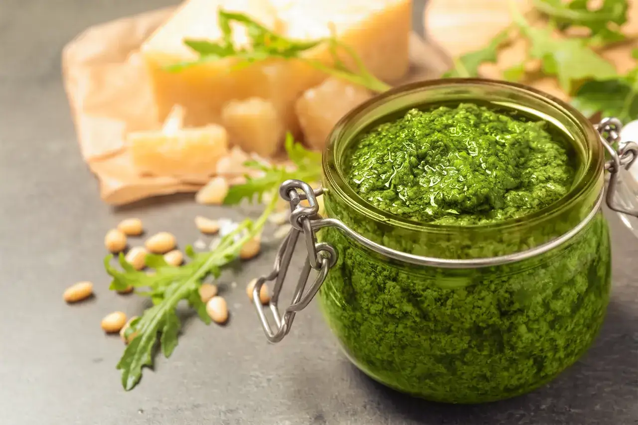 Pesto di rucola ricetta in un barattolo di vetro, con pinoli, rucola fresca e scaglie di Parmigiano sullo sfondo.