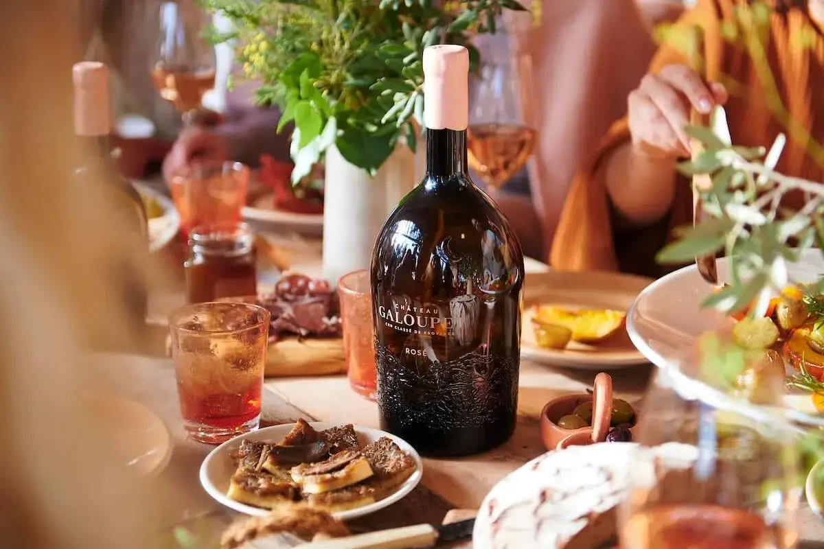 Un tavolino apparecchiato per un pasto all'aperto, con una bottiglia di rosato vino Chateau Galoupet al centro, contornata da stuzzichini e bicchieri.