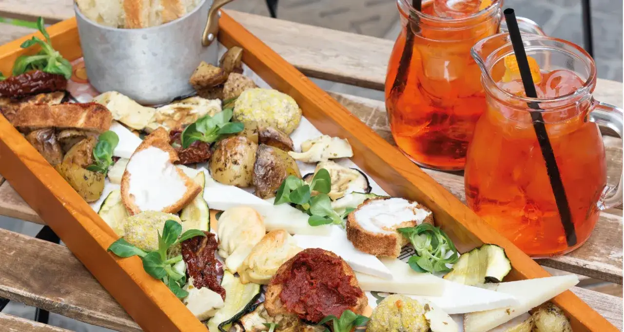 Un vassoio di stuzzichini con formaggio, pane, verdure grigliate e pomodori secchi, accompagnato da due bicchieri di spritz rinfrescante.