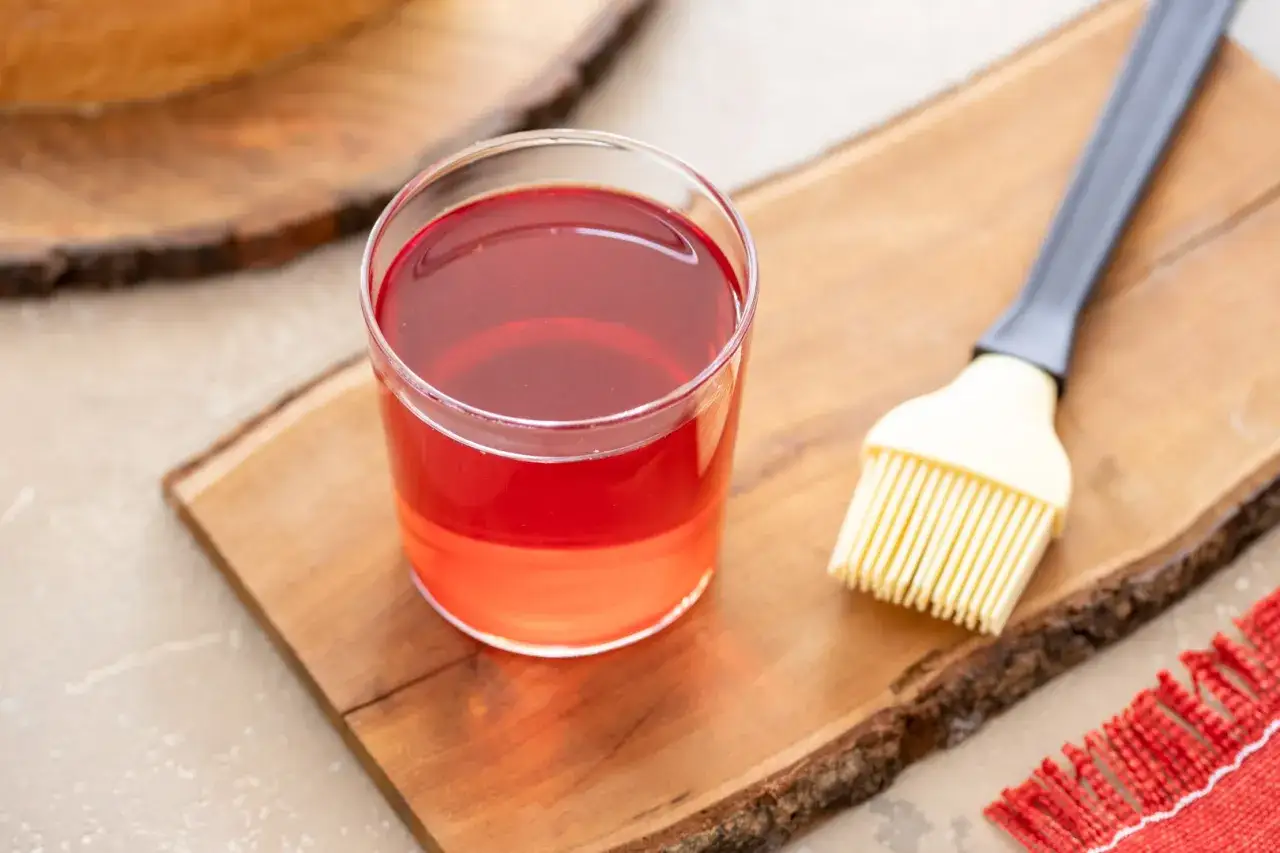 Bagna all'alchermes in un bicchiere, pronta per essere usata con un pennello da cucina su un tagliere di legno.