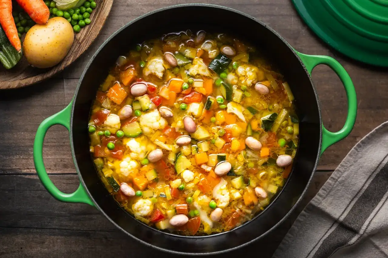 Minestrone di verdure fumante in una pentola verde, con carote, patate e cavolfiore visibili.