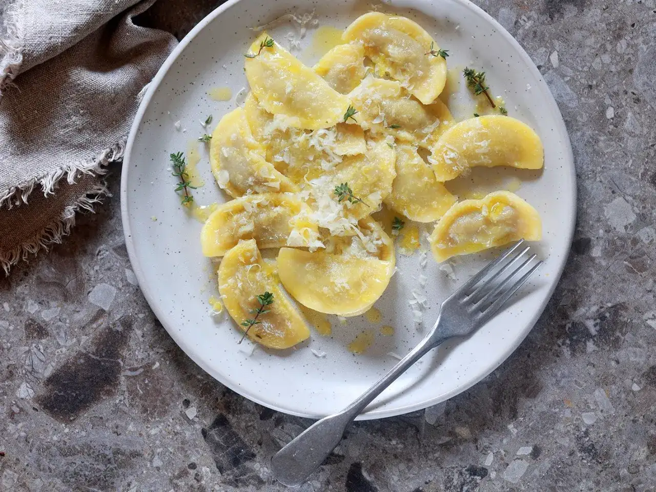 Piatto di tortelli cremaschi conditi con burro fuso, parmigiano grattugiato e rametti di timo. Una forchetta argentata &egrave; appoggiata sul bordo del piatto.