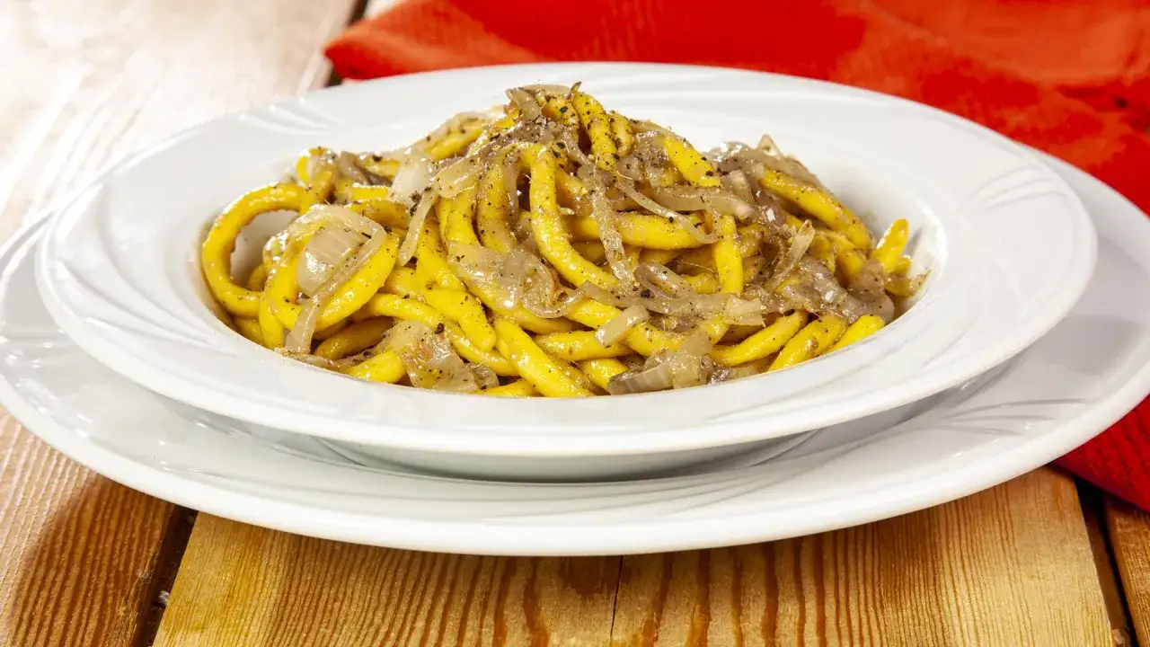 Un piatto di bigoli in salsa, pasta gialla condita con cipolle e pepe nero, su un tavolo di legno.