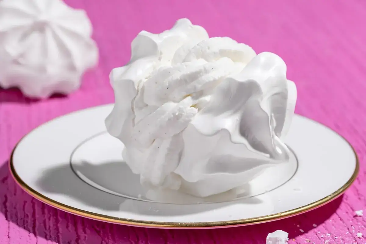 Dolce meringa soffice, simile a panna da montare, su un piatto bianco con bordo dorato, su sfondo rosa acceso.